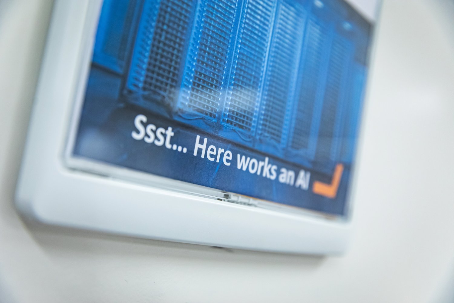 Close-up of a sign showing server racks with the text 'Ssst... Here works an AI' displayed in white letters on a blue background.